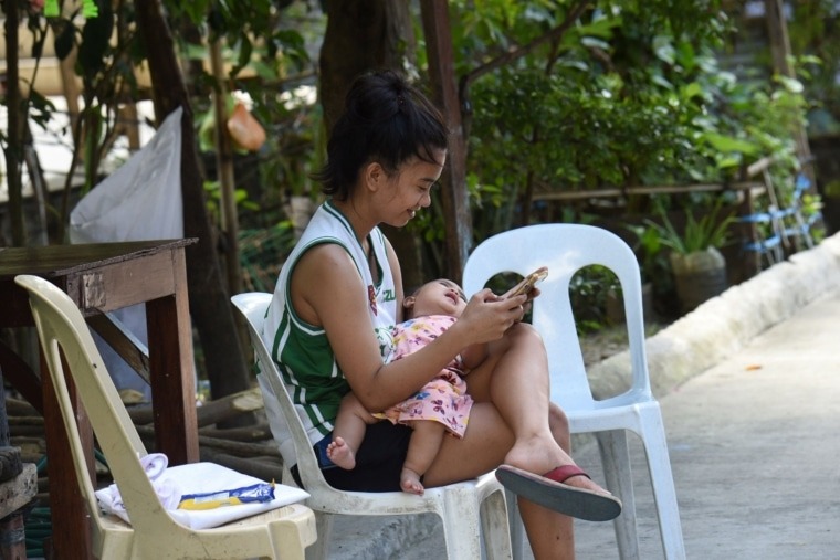 This photo taken on September 21, 2020 shows a teenage girl browsing her phone while holding a sibling's child as health officials conduct a polio vaccine campaign in the area, at a relocation site for informal settlers in San Jose Del Monte in Bulacan province, north of Manila. - Online misinformation is leaching out from cheap mobile phones and free Facebook plans used by millions of poor in the Philippines, convincing many to reject vaccinations for polio and other deadly diseases. (Photo by Ted ALJIBE / AFP) / TO GO WITH Health-virus-misinformation-internet-vaccine-Philippines,FOCUS by Jake Soriano (Photo by TED ALJIBE/AFP via Getty Images)